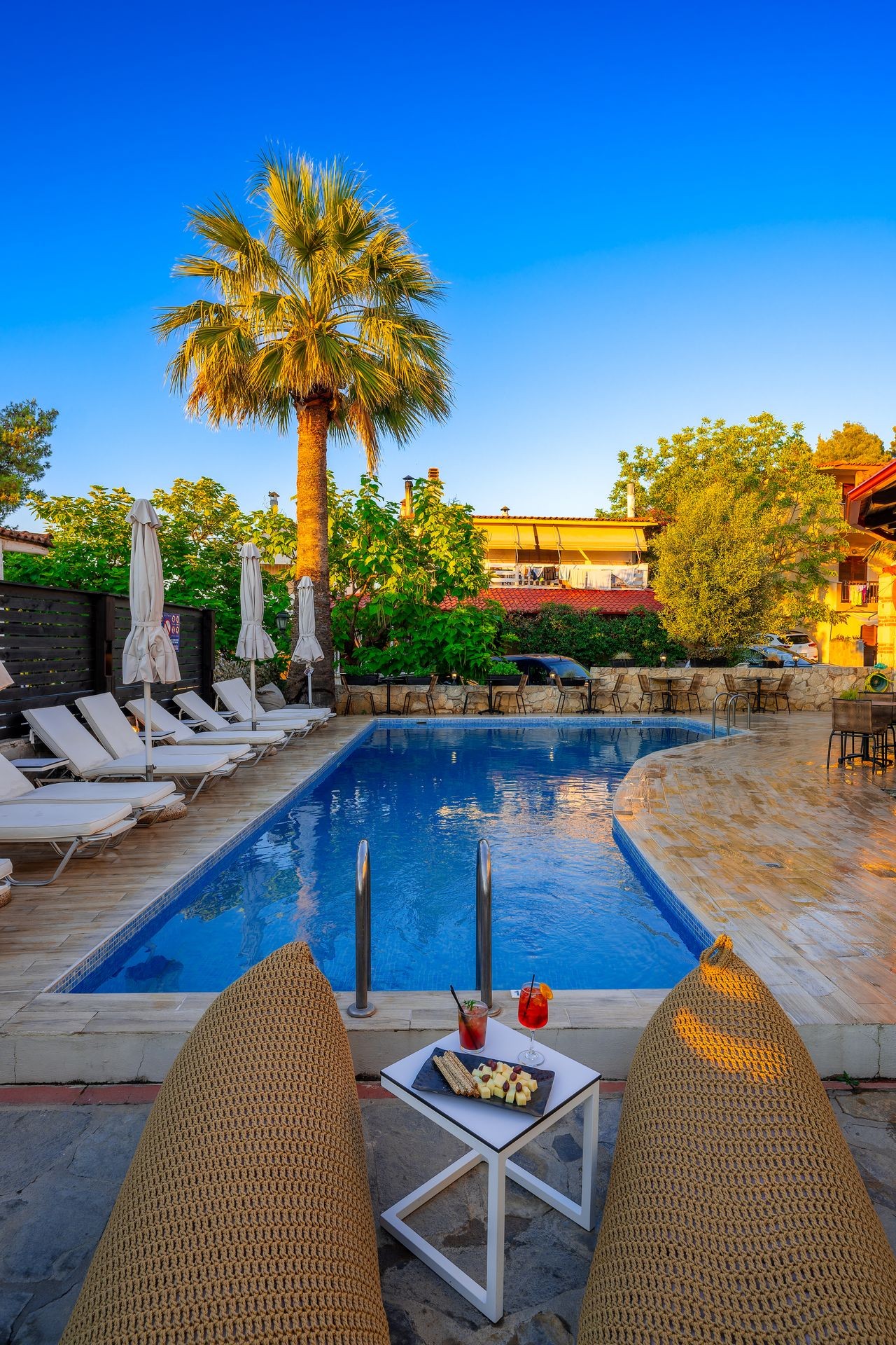 Outdoor pool area with lounge chairs, palm tree, and a small table with cocktails and snacks.